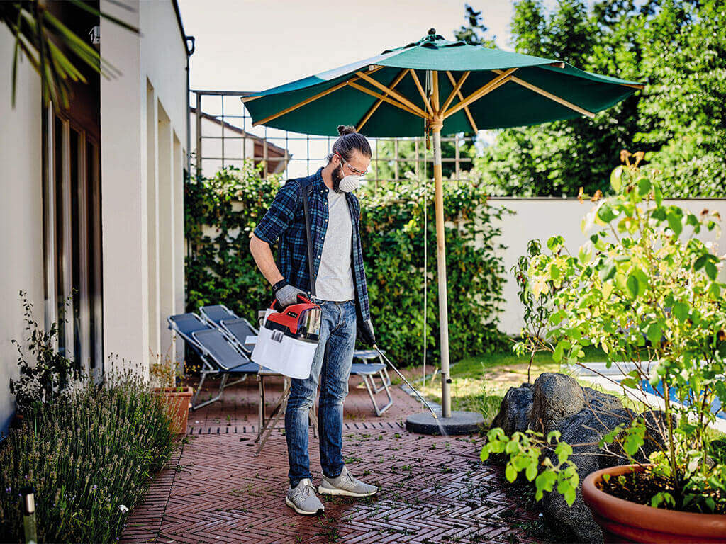 A man sprays pesticides on his plants in the garden with the help of an Einhell cordless pressure sprayer.