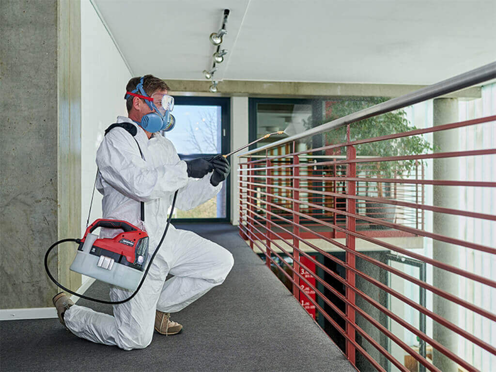 A man disinfects the handrail of a banister in the hallway with the help of an Einhell cordless pressure sprayer.