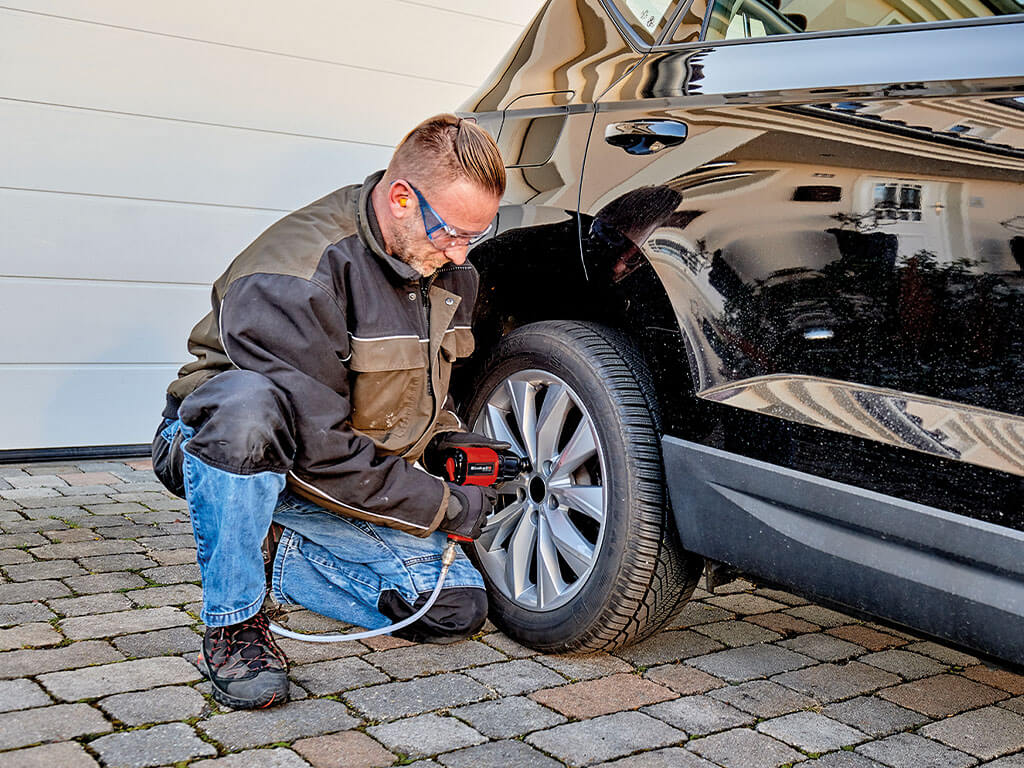 A man kneels next to a black car and loosens the wheel nuts of the tyre with an Einhell impact wrench.